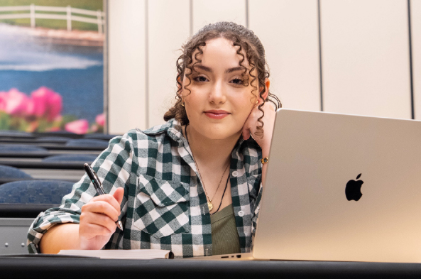 A student sits in a lecture hall behind her laptop while smiling.