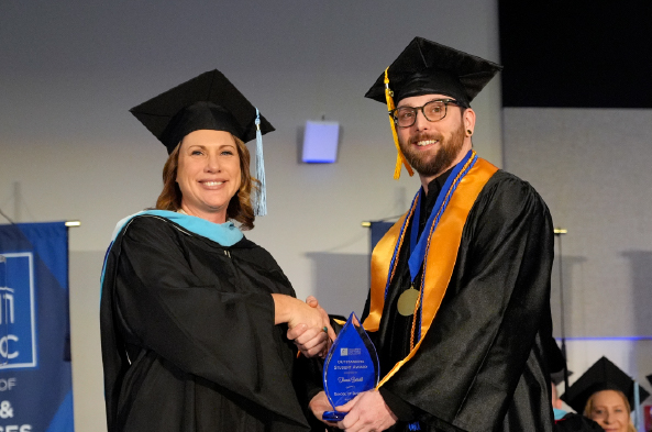 A graduating student shakes hands with a professor on the graduation stage. They are both wearing black regalia and smiling.