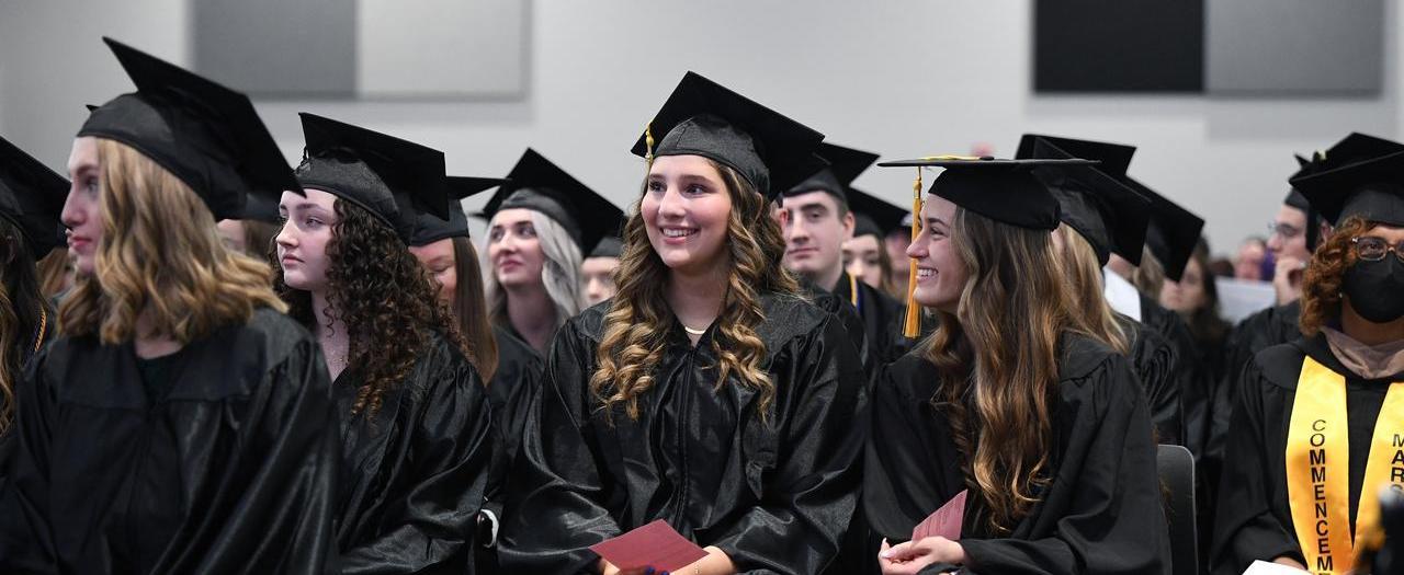 Graduates wearing black caps and gowns smile as they sit in an auditorium.