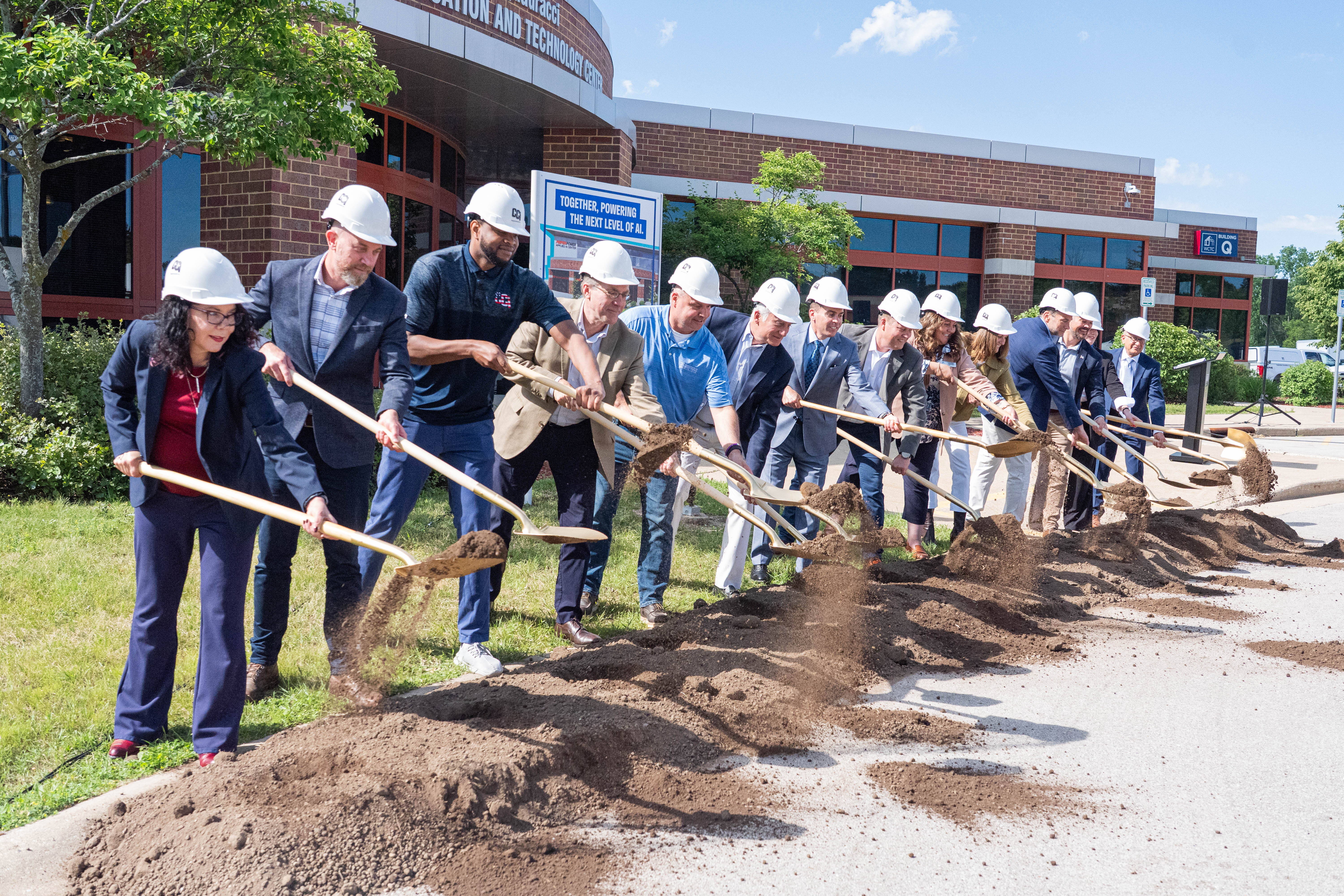 A line of WCTC leaders and community and business partners wearing hard hats all toss a shovelful of dirt at the same time