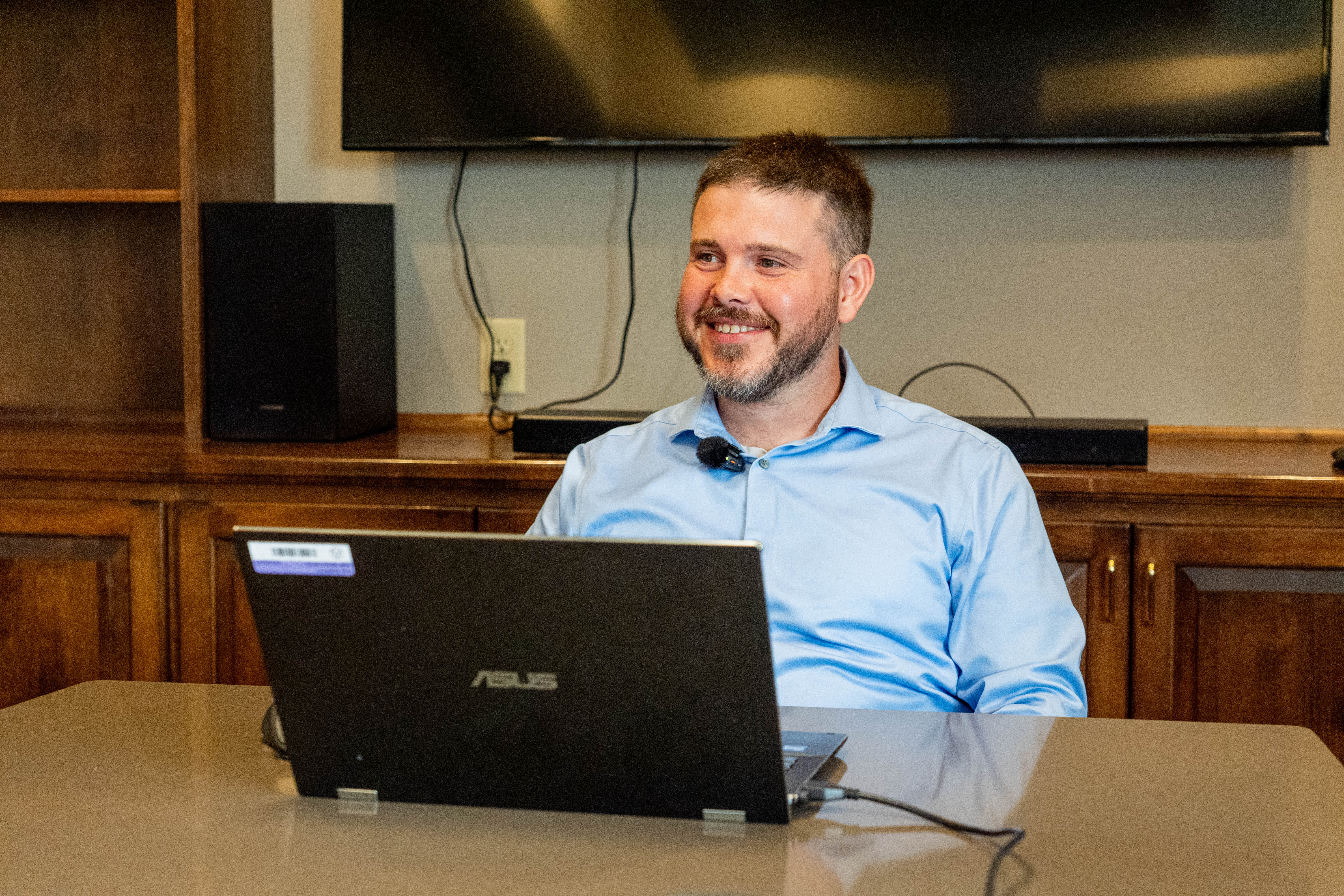 Alex Albrecht sits at a table smiling with a laptop open in front of him
