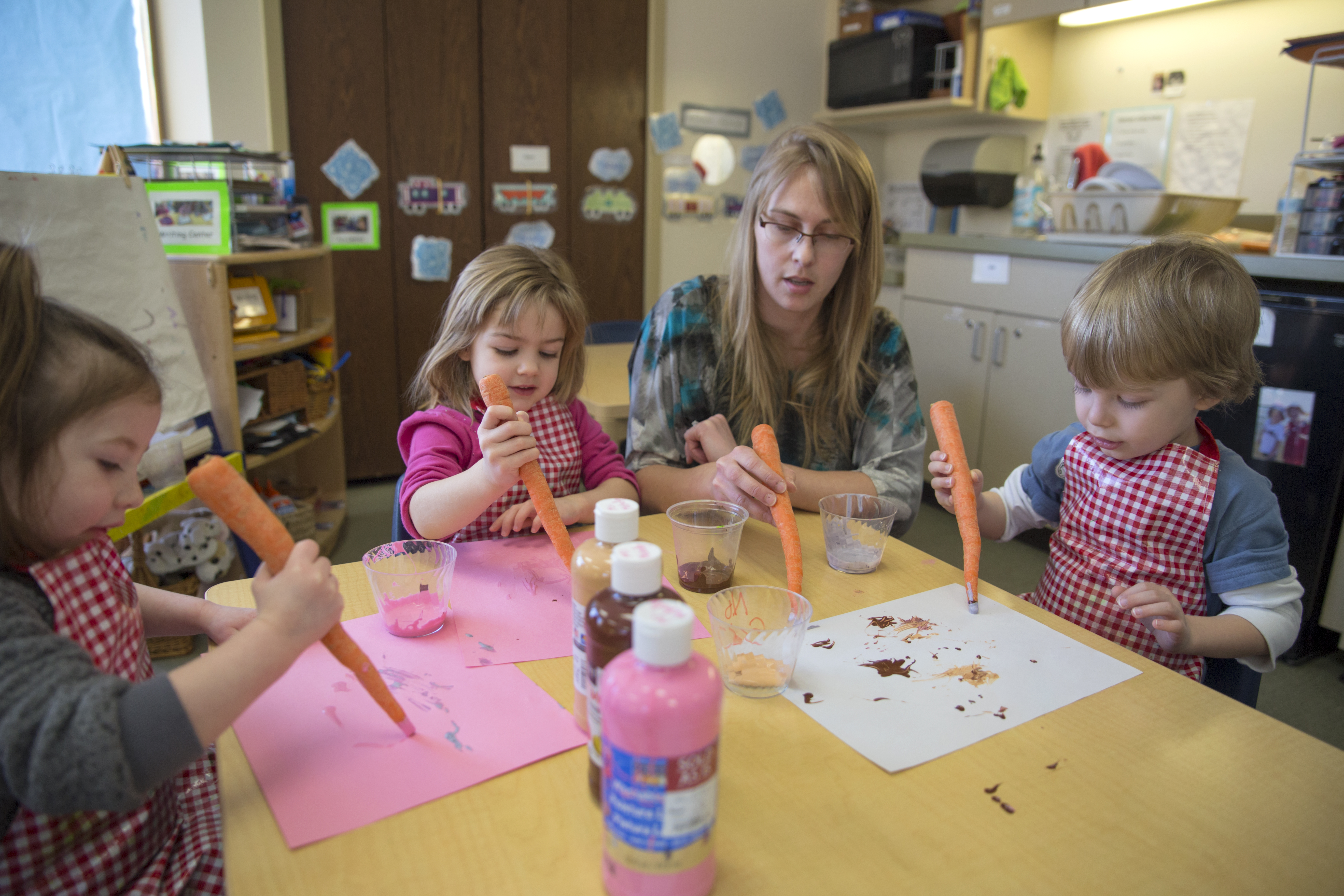 A young woman sits at a low table with three small children. They are all painting on paper using carrots as paintbrushes.