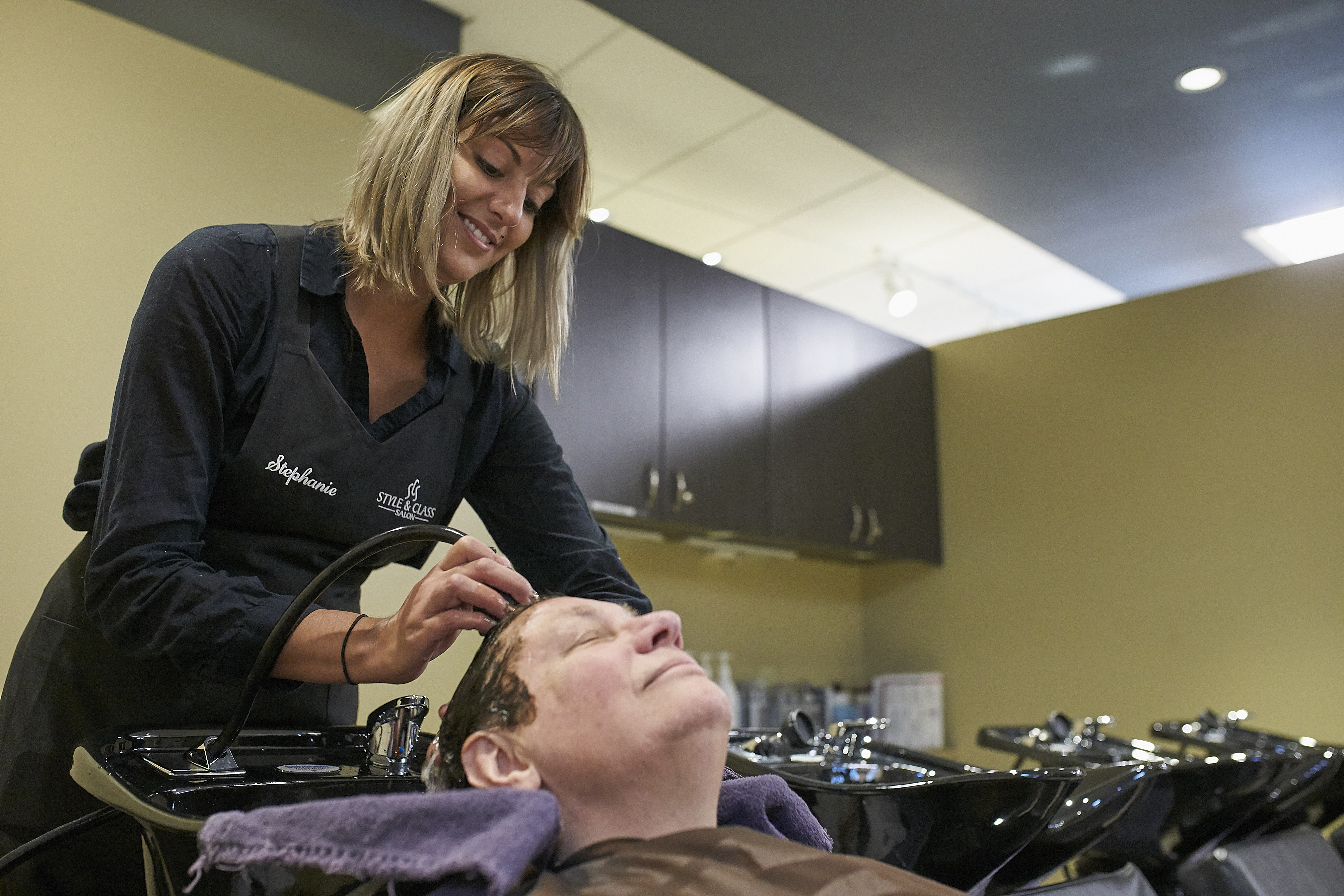 Cosmetology students in a black branded apron shampoos a woman's hair