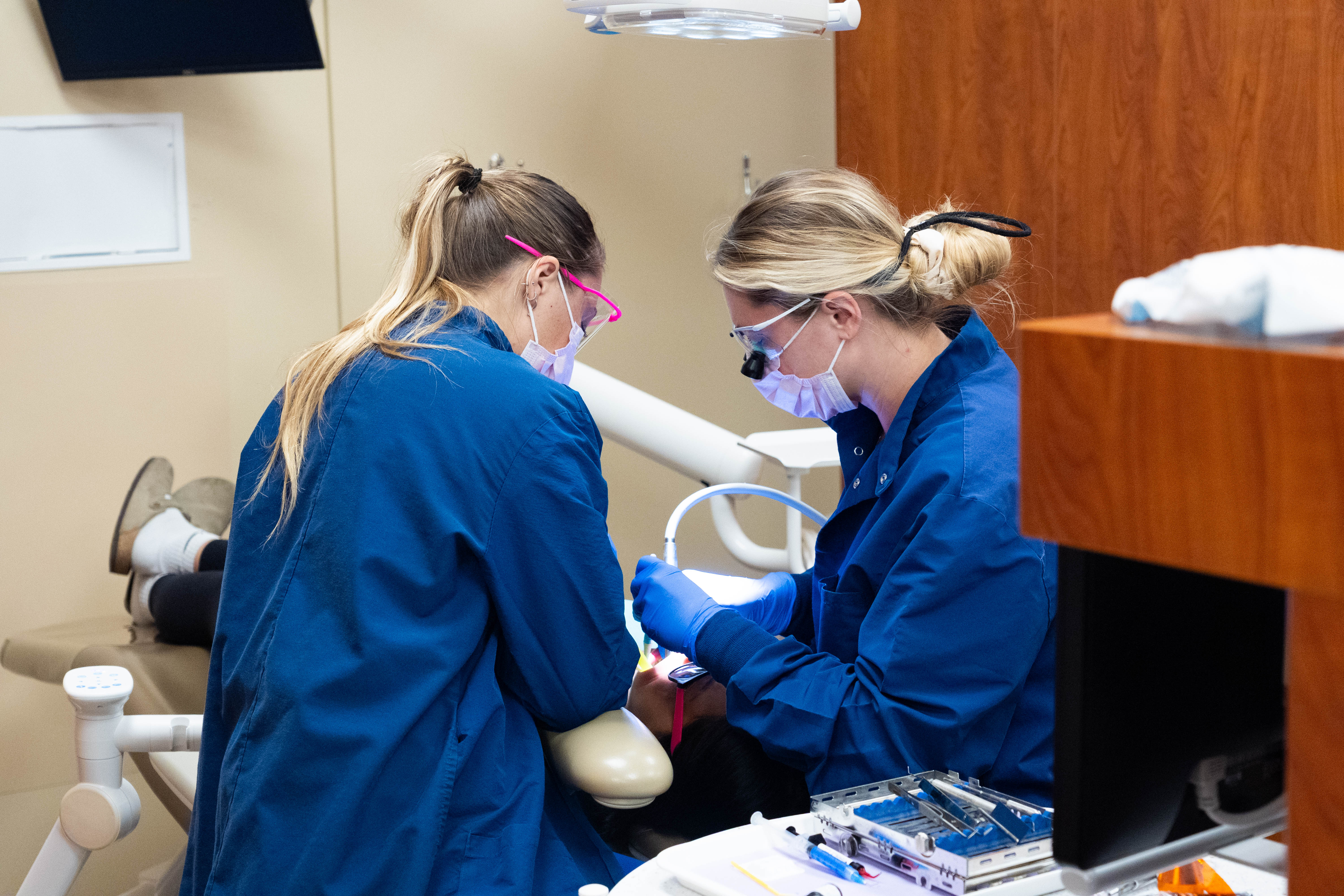 Two dental hygienist students in blue scrub coats work bend over working on a patient whose face you cannot see