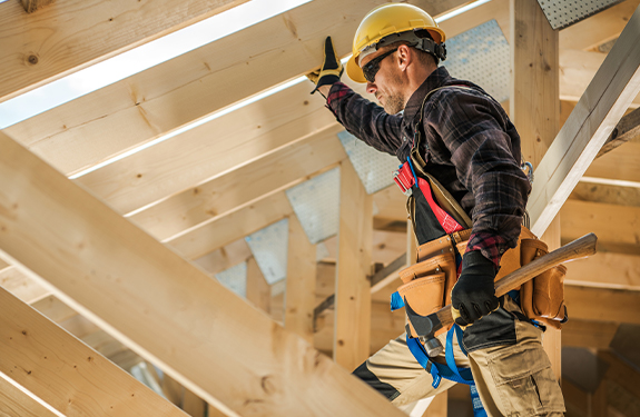 Man in a yellow hard hat with a hammer in his hand stands on a ladder considering some structural supports