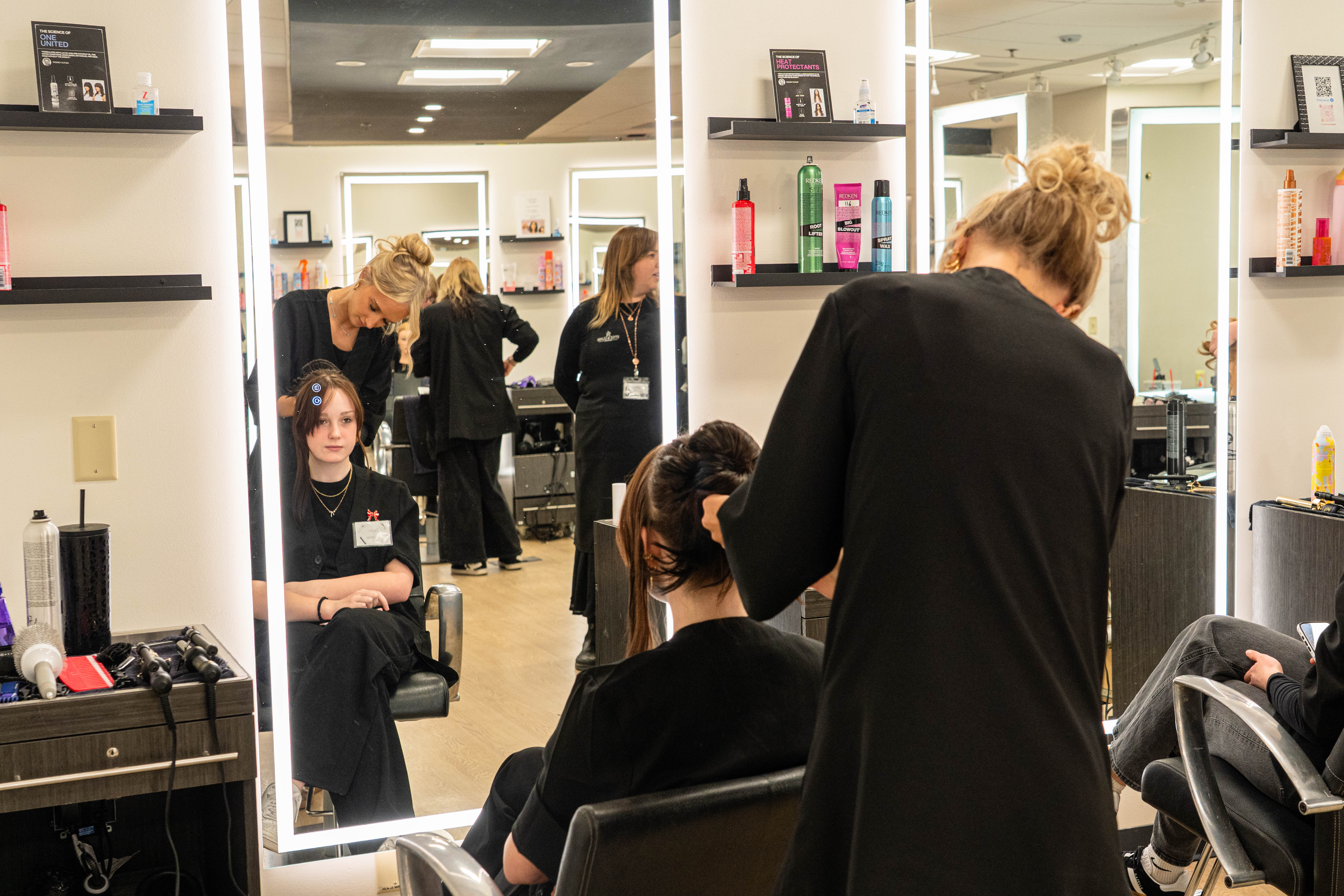A female student in black works on a woman's hair as she sits in a salon chair. You can see over their shoulders to their faces in the mirror.