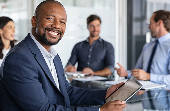 Man smiles at camera. He's holding a computer pad in a conference room with other people in the background talking